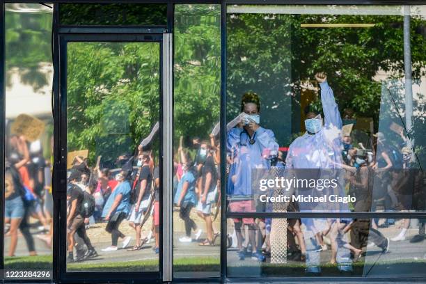 Medical professional at a plasma center holds her fist in the air as people marching to demand justice for Elijah McClain pass by the facility on...