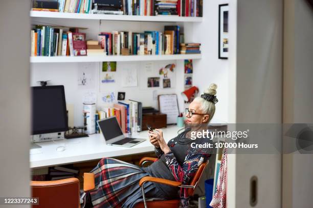 senior woman sitting in home office using phone - mid adult stock-fotos und bilder