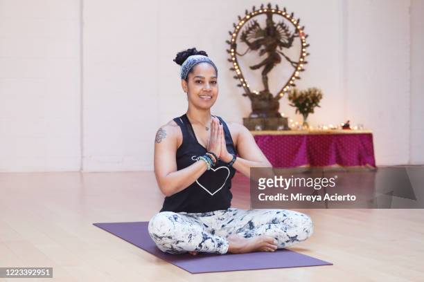 Portrait of a mature LatinX woman with curly hair, smiling with hands in prayer position, with a Hindu god sculpture in the background of a yoga studio.