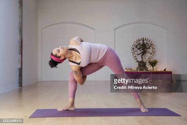 A mature LatinX woman with curly dark hair, stands in a bound extended side angle pose with a Hindu god sculpture in the background of a yoga studio.