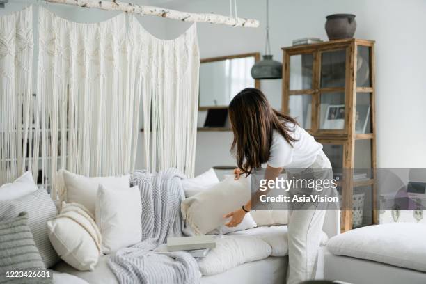 giovane donna asiatica che organizza e riordina i cuscini sul divano nel soggiorno di casa - arrangiare foto e immagini stock