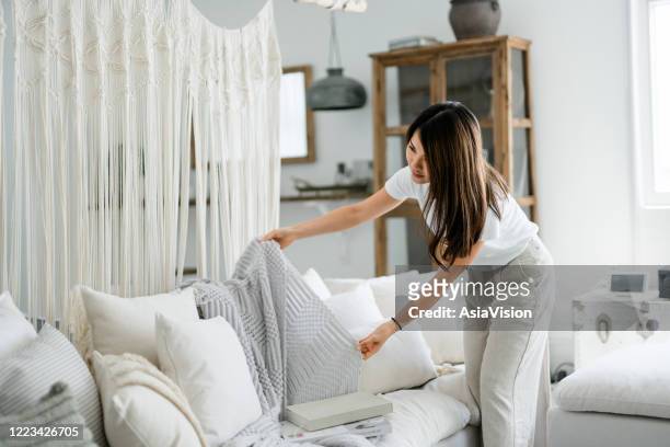 young asian woman organising and tidying up the cushions and throw on the sofa in the living room at home - home interior stock pictures, royalty-free photos & images