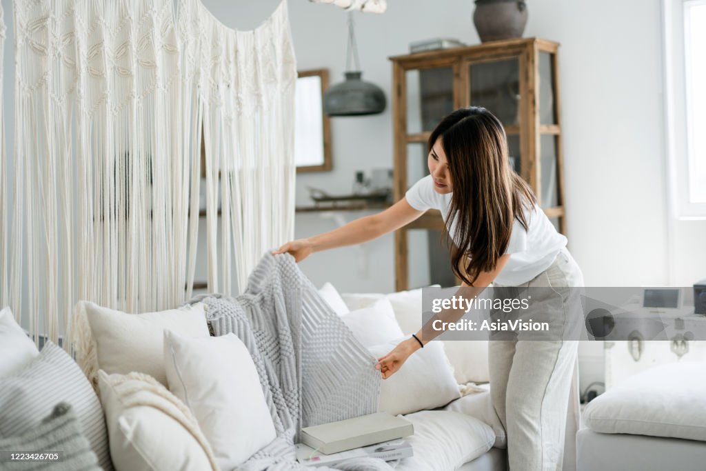 Young Asian woman organising and tidying up the cushions and throw on the sofa in the living room at home