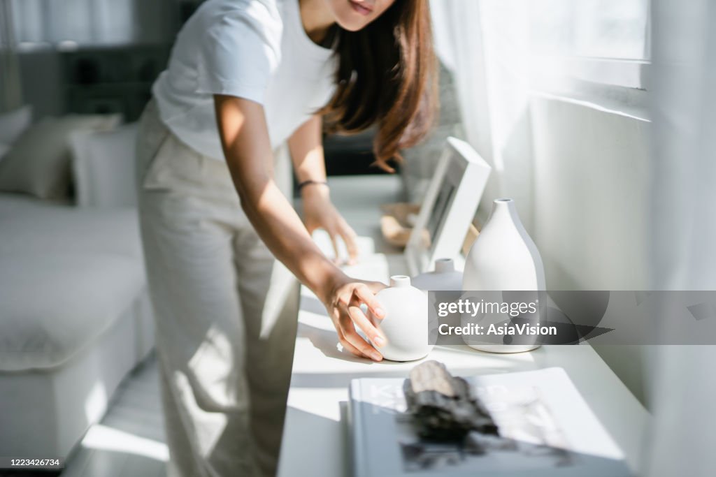 Jovem asiática aproveita seu tempo em casa, decorando e organizando quadros e vasos na prateleira perto da janela