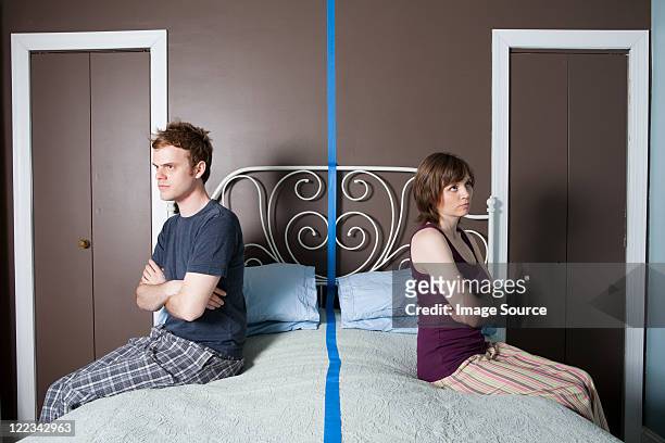 young couple sitting on bed separated by blue line - stubborn stock-fotos und bilder