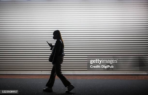 a young woman wearing a mask operates her smartphone while walking, silhouetted against closed shutter in the night - evening walk stock pictures, royalty-free photos & images