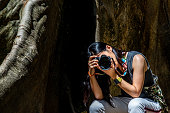 Young woman with Shoulder bag and using a camera to take photo Giant big tree, Size comparison between human and giant big tree in Ban Sanam of Uthai Thani Province, Thailand.