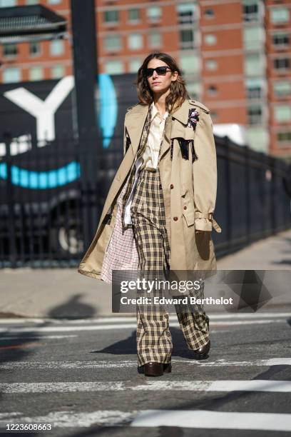 Guest wears sunglasses, a beige oversized trench coat with checked inner lining, checked flared flowing pants, brown leather shoes, a shirt, a...