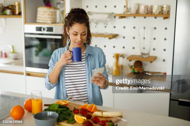 young woman using her mobile phone while preparing fruit smoothie in the kitchen - mixing stock pictures, royalty-free photos & images