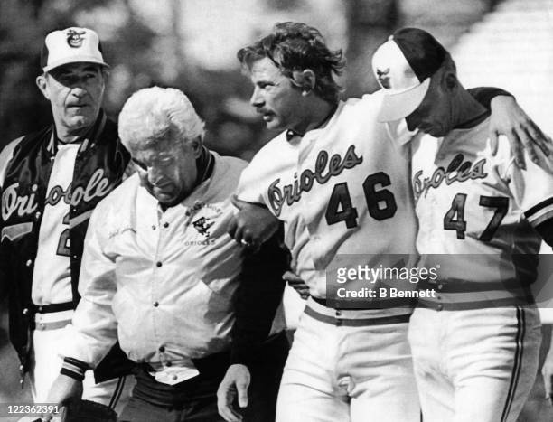 Pitcher Mike Flanagan of the Baltimore Orioles is helped off the field by manager Joe Altobelli, trainer Ralph Salvone and Cal Ripken Sr. #47 after...