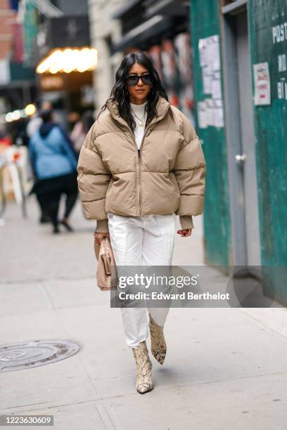 Guest wears sunglasses, a white turtleneck pullover, a pale pastel brown oversized puffer coat, white pants, snake print pointy leather boots, a bag,...