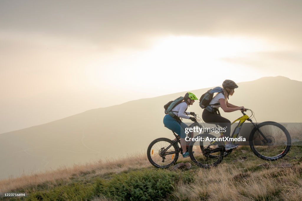 Two women ride up grassy hillside on electric mountain bikes