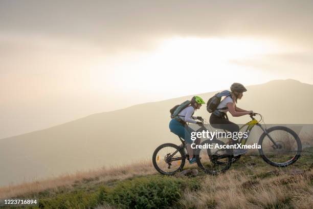 two women ride up grassy hillside on electric mountain bikes - e bike stock pictures, royalty-free photos & images