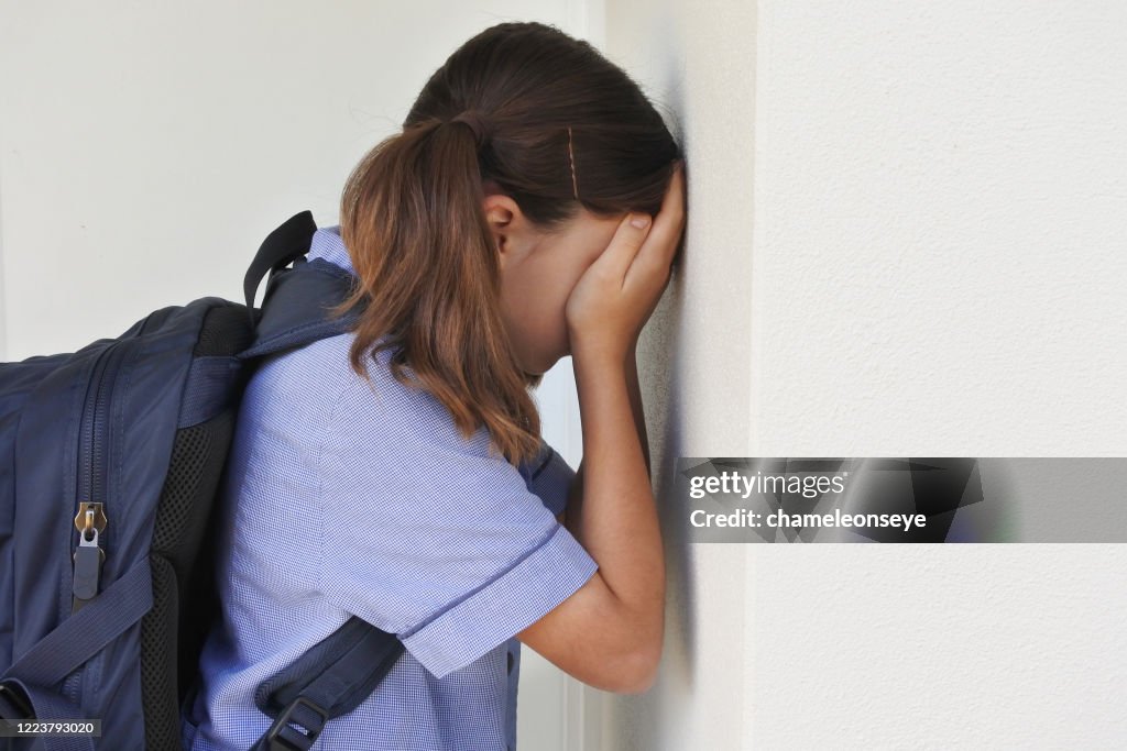 Triste joven colegiala cubriéndose la cara y llorando contra una pared