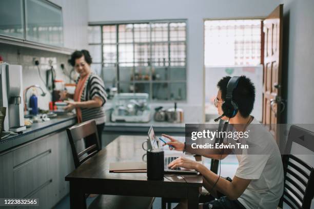 un adolescent chinois asiatique bavardant en ligne avec tous ses camarades de classe par l’intermédiaire du logiciel en ligne tout en faisant ses devoirs à la table à manger de cuisine tandis que sa grand-mère cuisinant dans la cuisine préparant la  - teenage boys stock photos et images de collection