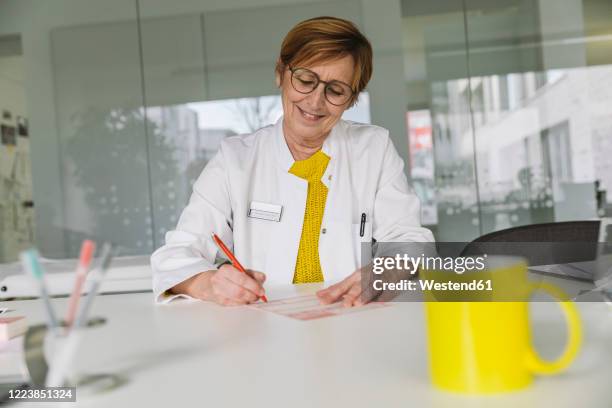 doctor sitting at desk filling out prescription - verschreibungspflichtiges medikament stock-fotos und bilder