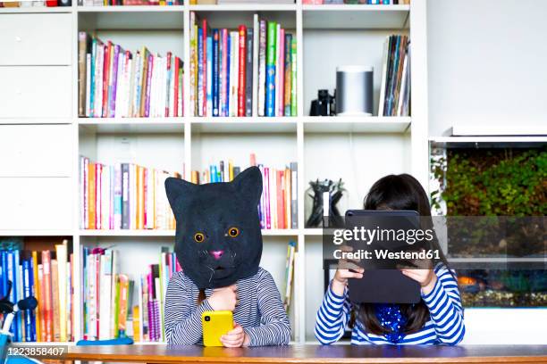 two girls sitting at table, hiding behind digital tablet and cat mask - förklädnad bildbanksfoton och bilder