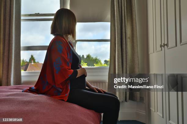 lone pregnant woman sitting on the bed looking out at the window in the morning - covid separation stock pictures, royalty-free photos & images