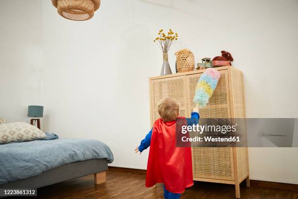 boy wearing superman costume and cleaning with feather duster on a sideboard at home - anrichte stock-fotos und bilder