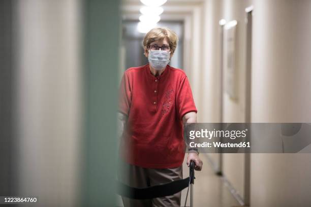 senior woman wearing mask and walking with wheeled walker in corridor of retirement home - rollator stock-fotos und bilder