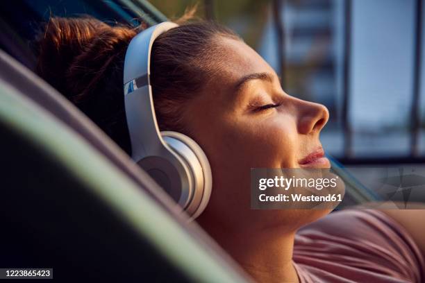 smiling young woman lying on hammock on balcony listening music with headphones - music home stock-fotos und bilder