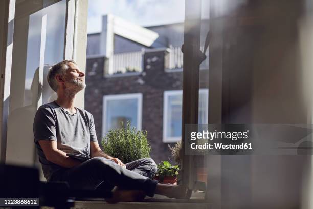 mature man sitting at the window at home with closed eyes - tranquilidad fotografías e imágenes de stock