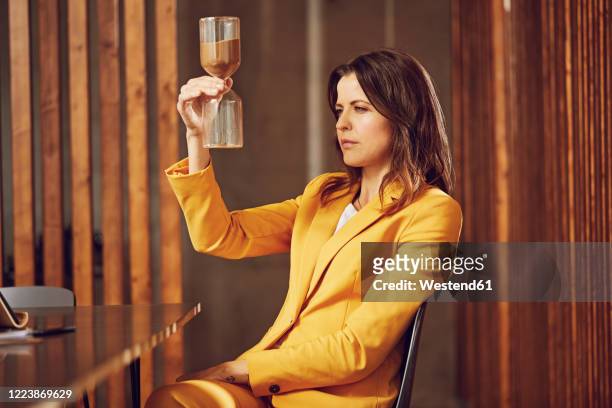 businesswoman wearing yellow suit sitting at desk in office holding hourglass - running water stock-fotos und bilder
