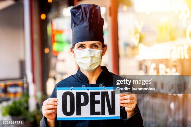 female chef restaurant owner posing wearing a mask holding an open sign - restaurants open during lockdown stock pictures, royalty-free photos & images