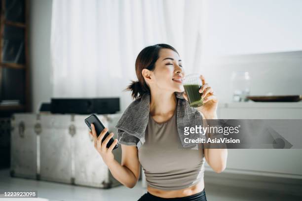 jeune femme asiatique de sports se reposant et ayant le smoothie vert après l’exercice à la maison le matin, utilisant le smartphone - asian drink photos et images de collection