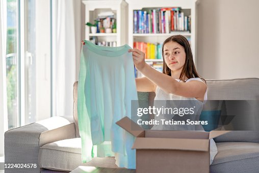 Disappointed young teenager girl unpacking parcel with garment delivered in box by courier to house looking at dress of wrong size. Online shopping concept. Dissatisfied with online purchase or gift Disappointed young teenager girl unpacking parcel with garment delivered in box by courier to house looking at dress of wrong size. Online shopping concept. Dissatisfied with online purchase or gift