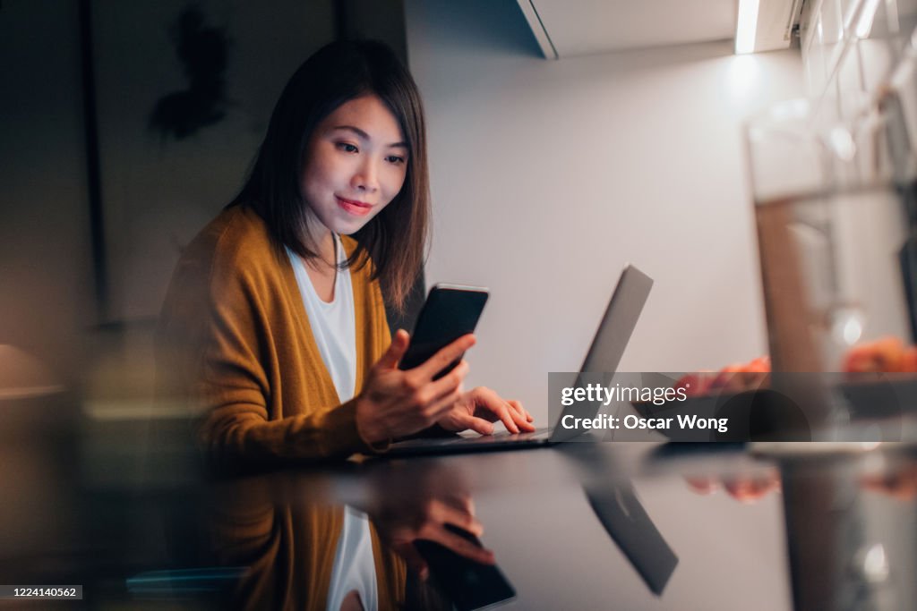 Young Woman Using Smartphone While Working With Laptop At Home In The Dark