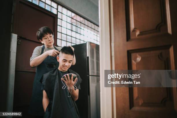 an asian chinese mid adult woman is cutting and trimming hair for her son in the kitchen due to travel ban - teenage boy shave imagens e fotografias de stock