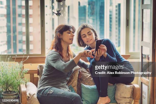 a mother and daughter sharing a mobile phone in their apartment - mother and teenage daughter foto e immagini stock