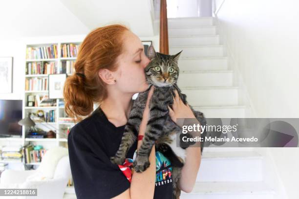 teenage girl with red hair holding her pet cat and giving it a kiss. she is in her house standing at the bottom of a staircase. the living room can be seen behind her and the house is painted all white. - girls cuddling cat stock-fotos und bilder
