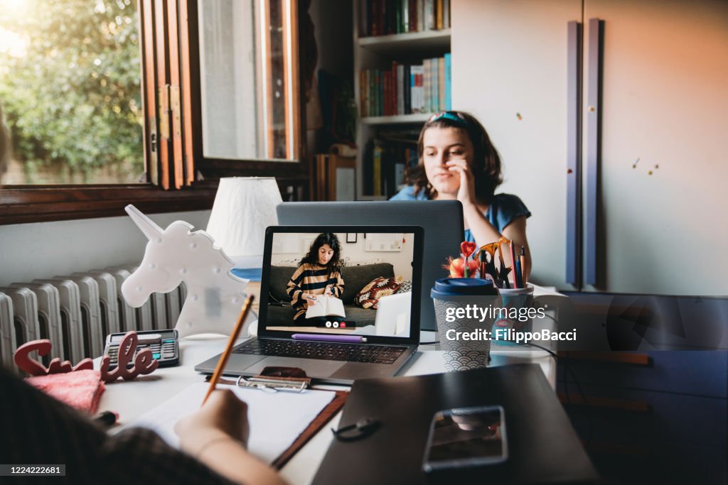 Two sisters are studying together at home, sharing a desk to participate at online school lessons with laptops