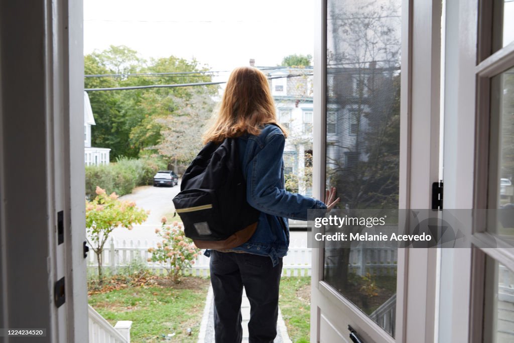 Teenage girl walking out the front door of her house. Back view of her leaving the house. She is on her way to school, wearing a back pack and holding the door open.