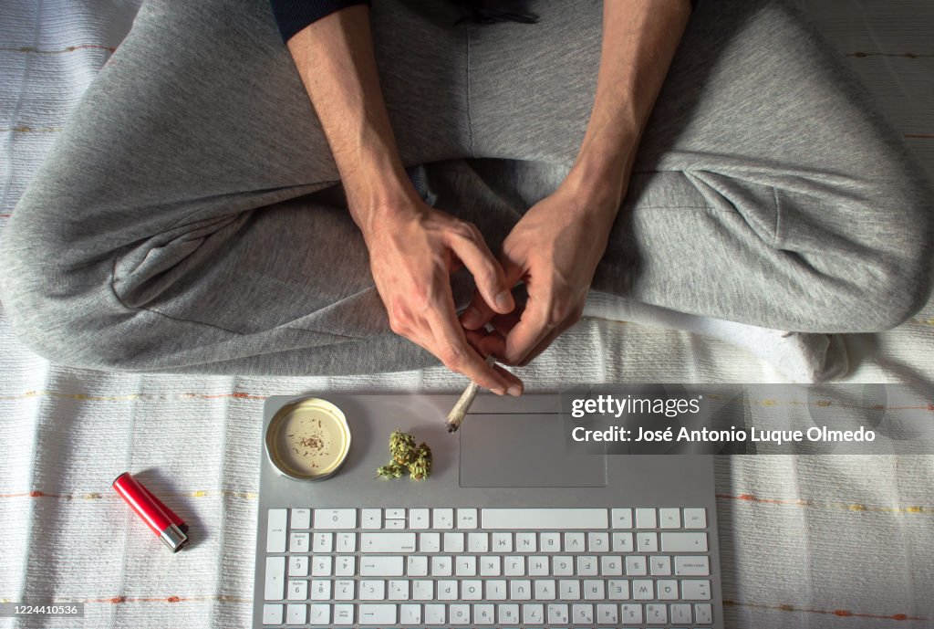 Vista superior de la persona sentada en una cama fumando marihuana mientras usa un ordenador portátil para ver un video o película. Concepto de cannabis y tecnología.