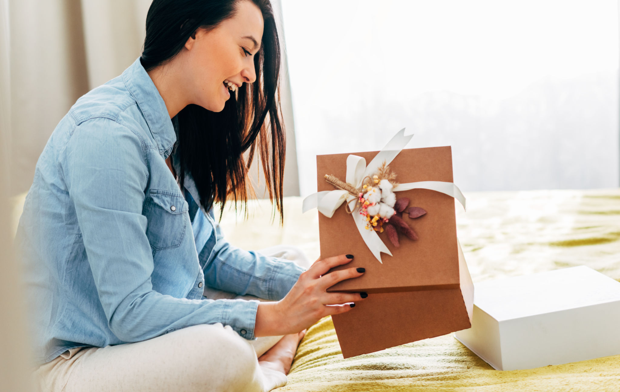 Side view image of happy young woman sit on the bed unpacking the parcel received. Pretty female holding gift box receive order from online shop purchase at home. Online internet commerce delivery Side view image of happy young woman sit on the bed unpacking the parcel received. Pretty female holding gift box receive order from online shop purchase at home. Online internet commerce delivery