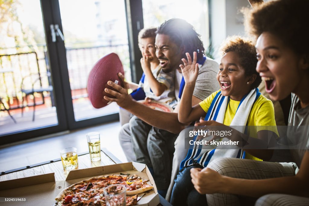 Família negra alegre torcendo enquanto assiste jogo de rúgbi na TV em casa.
