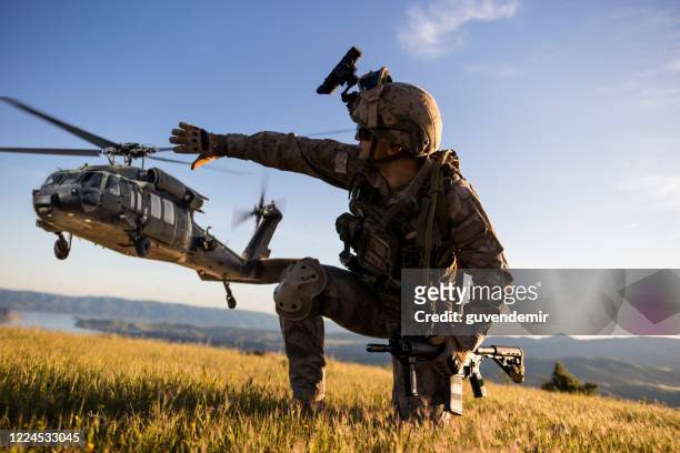 military helicopter approaching behind the kneeling army soldier - tropa imagens e fotografias de stock