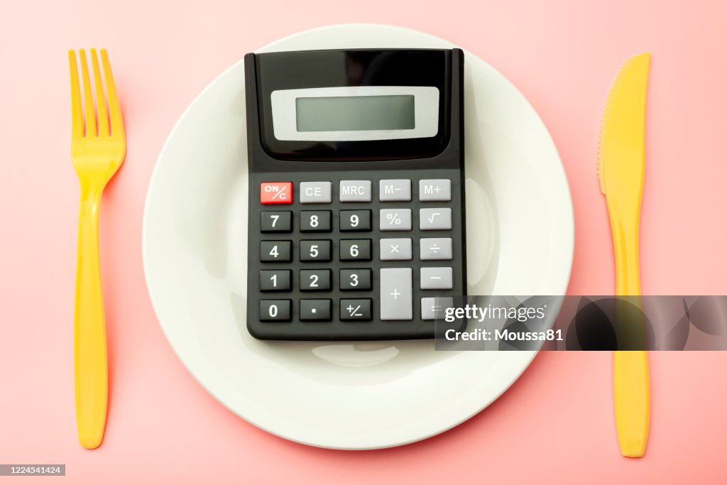 Calculate expensive food spending costs, counting calories and weight loss program concept with calculator on empty plate, yellow fork and knife isolated on pink background