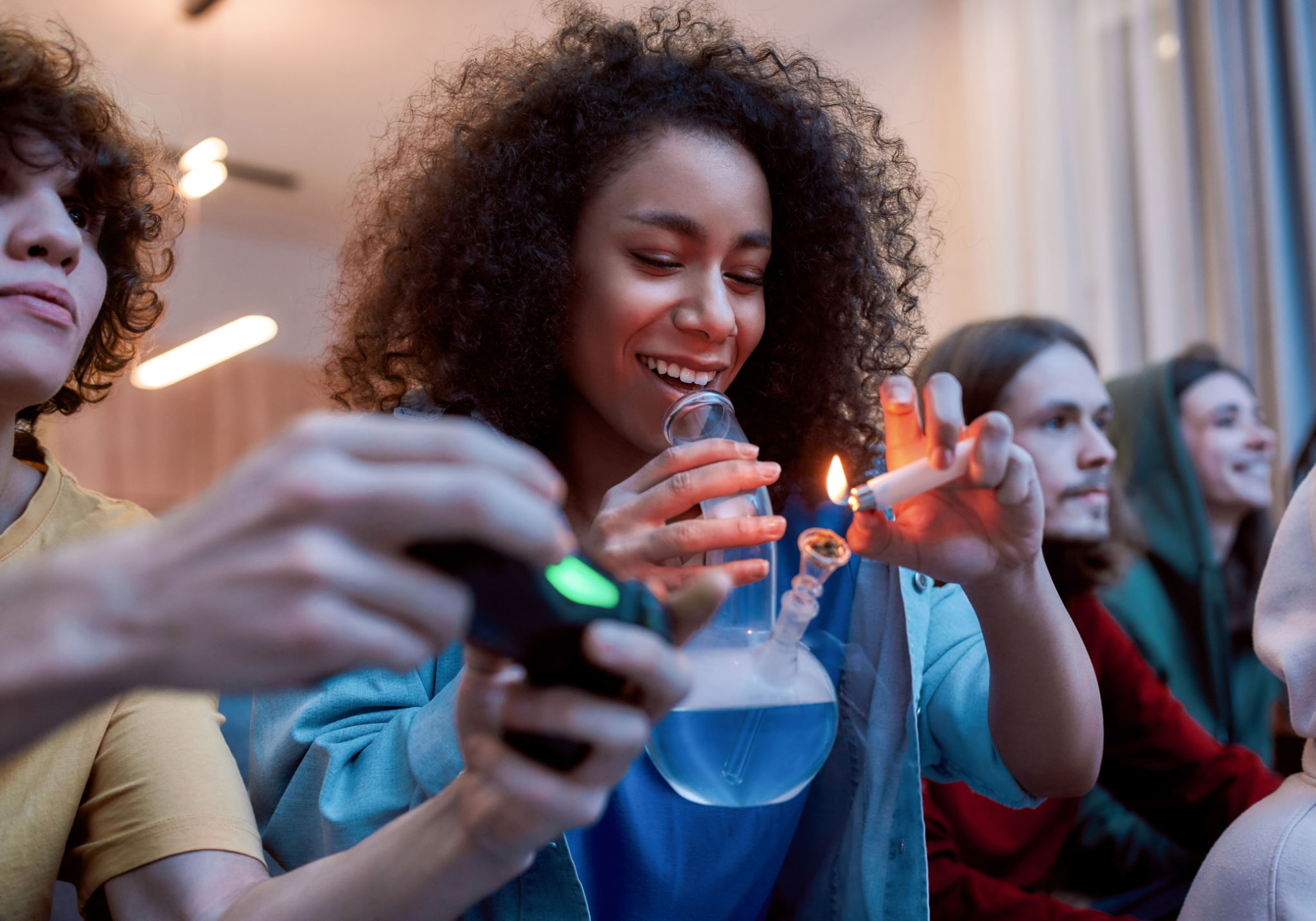 For great mood. Young afro american girl lighting marijuana in the glass bong, relaxing with friends on the sofa at home. Young people playing video games and smoking weed For great mood. Young afro american girl lighting marijuana in the glass bong, relaxing with friends on the sofa at home. Young people playing video games and smoking weed