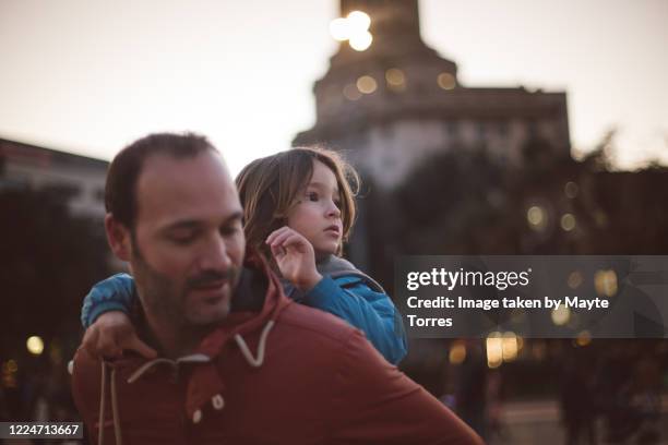 dad carrying boy on his back in the city while he looks away - neue männlichkeit stock-fotos und bilder