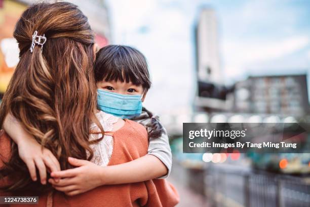back view of young mom carrying her tired little daughter in medical face mask while strolling in city street - luchtvervuiling stockfoto's en -beelden