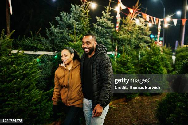 smiling couple walking through christmas tree lot while shopping for tree - winter coat stock pictures, royalty-free photos & images