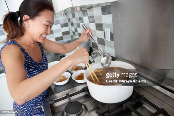 an asian woman wearing a ponytail and summer dress, smiles while stirring homemade soup with chopsticks and a large spoon in her home. - chinese soup photos et images de collection