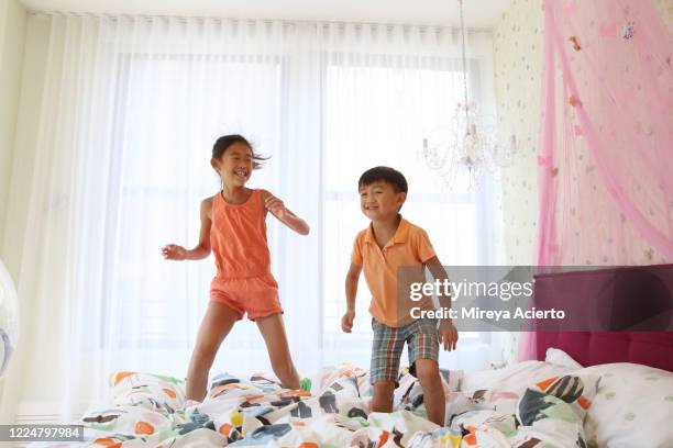 two young asian children wearing summer clothes, jump on the bed in a colorful, brightly lit bedroom. - a boy jumping on a bed fotografías e imágenes de stock