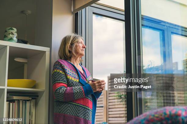 senior woman looking out of window with a cuppa - quarantäne stock-fotos und bilder