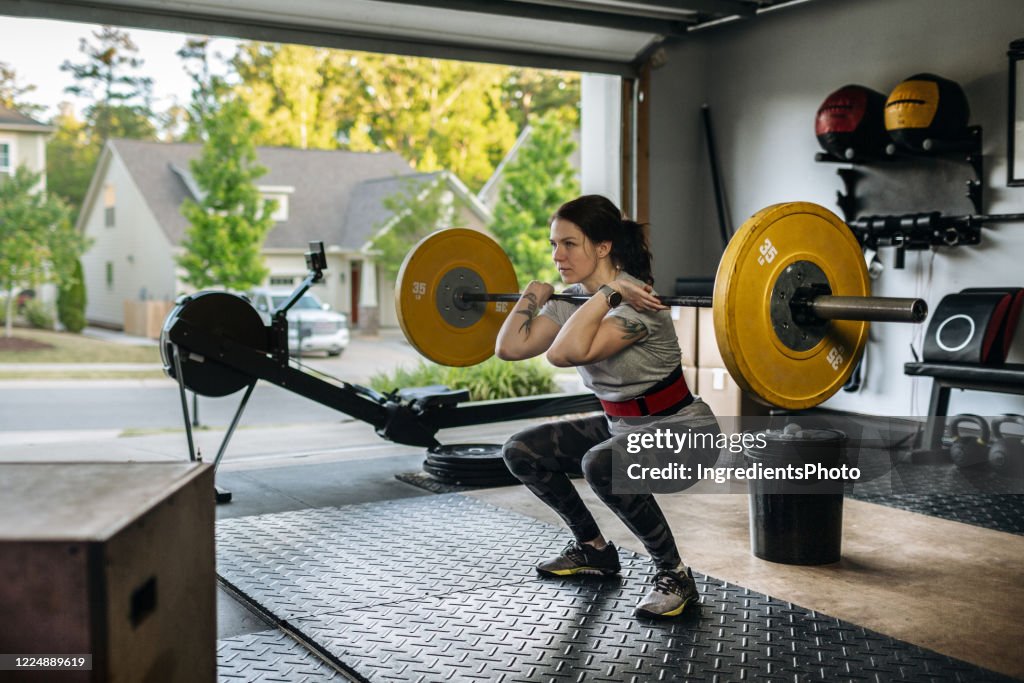 Fit mujer realizando sentadilla delantera con barbell pesado en el gimnasio de su casa en el garaje durante la pandemia de Covid-19.