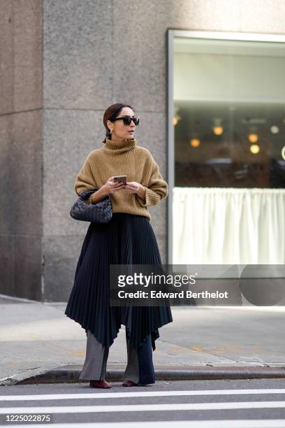 Geraldine Boublil wears sunglasses, a brown wool turtleneck pullover, a black woven leather Bottega Veneta bag, a dark blue pleated skirt, gray...
